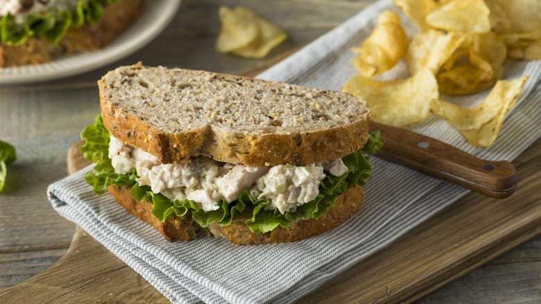 Chicken salad sandwich on multigrain bread on wooden cutting board with potato chips