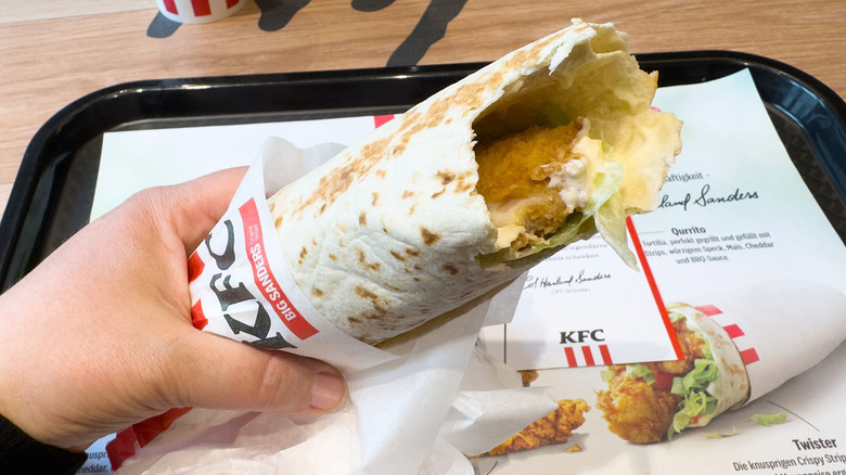 Hand holding KFC fried chicken wrap in wrapper above tray at restaurant