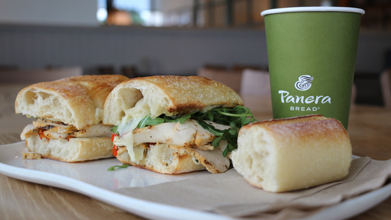 Tuscan Chicken Sandwich split in half on plate with side of baguette bread next to Panera takeout coffee cup in restaurant
