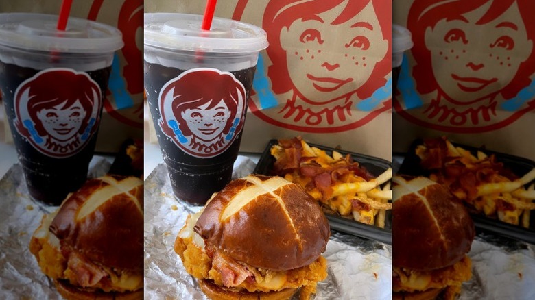 The Pretzel Bacon Pub Chicken Sandwich from Wendy's on foil next to takeout soda, bacon-topped fries, and Wendy's takeout bag