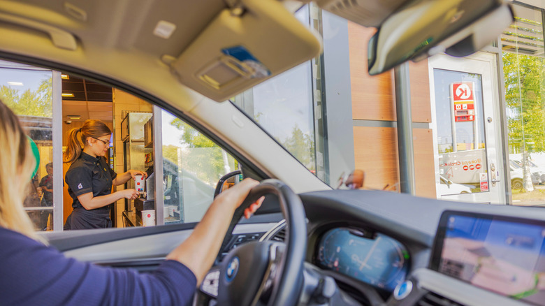 A drive-thru window picture taken from inside a car with woman in car looking at fast food worker preparing sodas