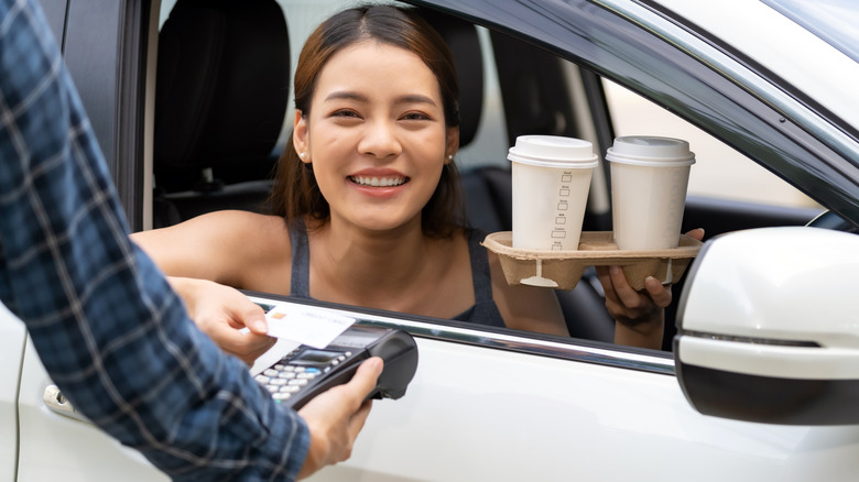 A woman smiling while paying for two coffees in to-go holder at drive-thru, holding credit card to card reader