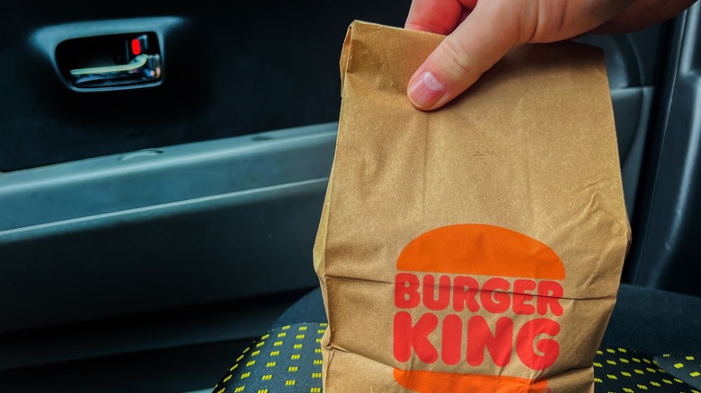 A hand holds the top of a Burger King paper bag in the front passenger's seat of a car