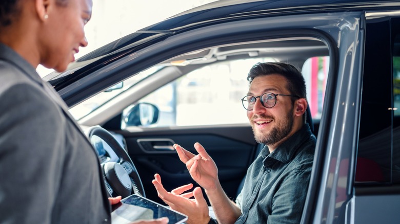 A woman in a suit and with a tablet approaches a man smiling in his car as he gestures and explains