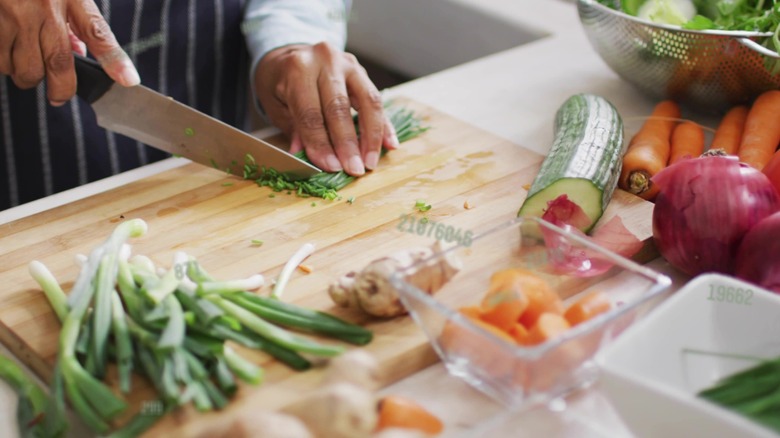 Hands seen chopping various items on wooden board in kitchen, wearing apron, using chef's knife. Closeup, slicing, cooking, home, produce, vegetables, natural
