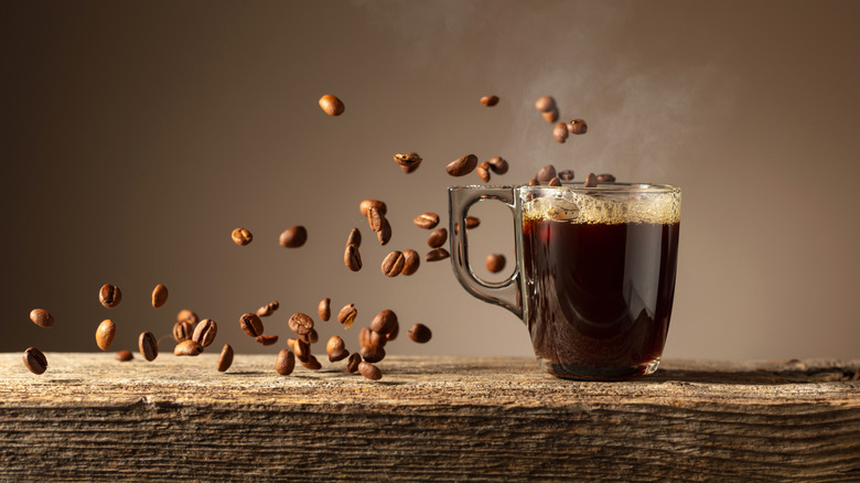 A glass cup of coffee on a rustic wooden table with coffee beans falling around it.