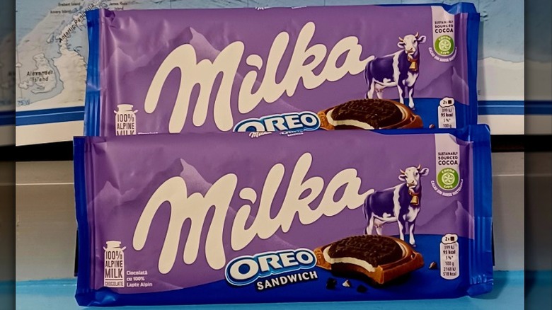 Two bags of Milka Oreo sandwich chocolate bars with purple and blue packaging stacked on a blue surface in front of a map