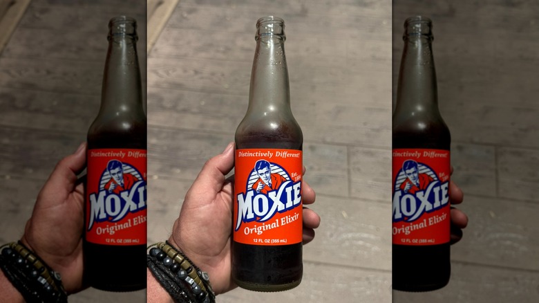 A hand holding an open glass bottle of Moxie soda