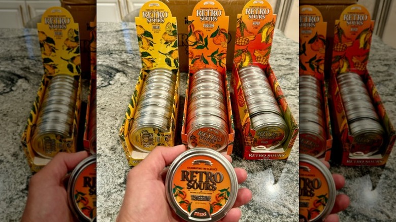 A hand holding a tin of Iconic Candy's Retro Sours in front of tins of citrus, tangerine, and mango flavors