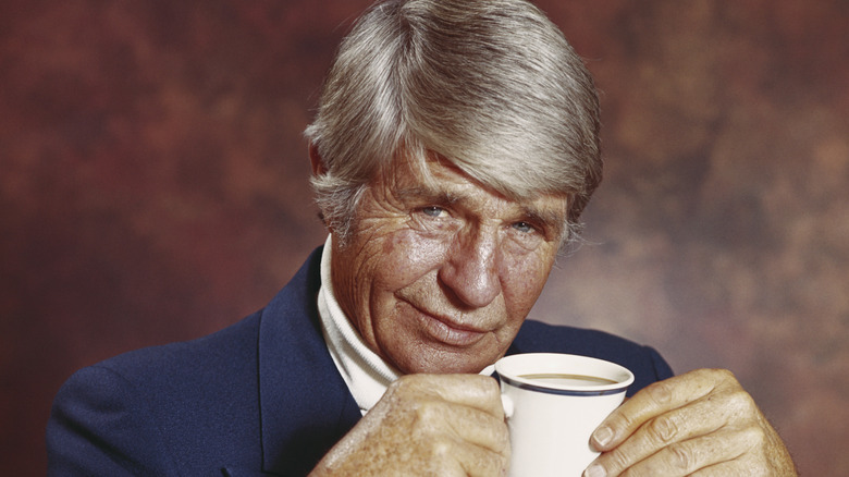 A older man from the 1970s holding a mug and smirking at camera