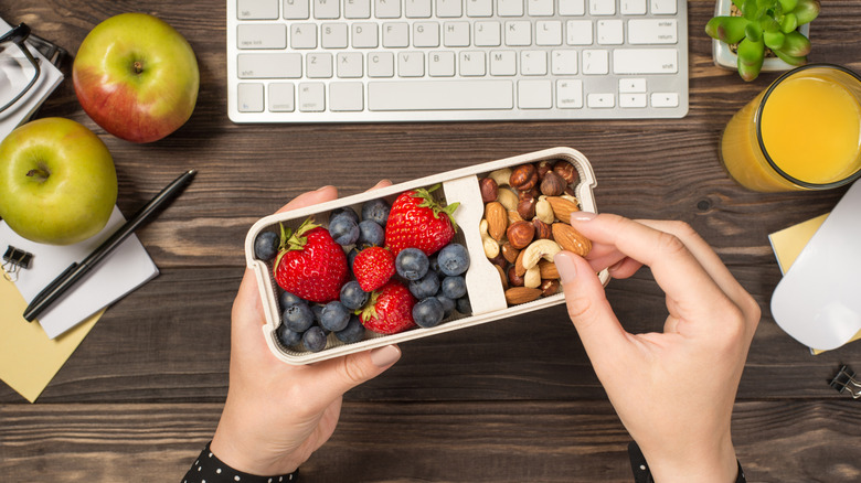 A person grabs a healthy snack of berries and mixed nuts at her desk during work with a glass and two apples on the side