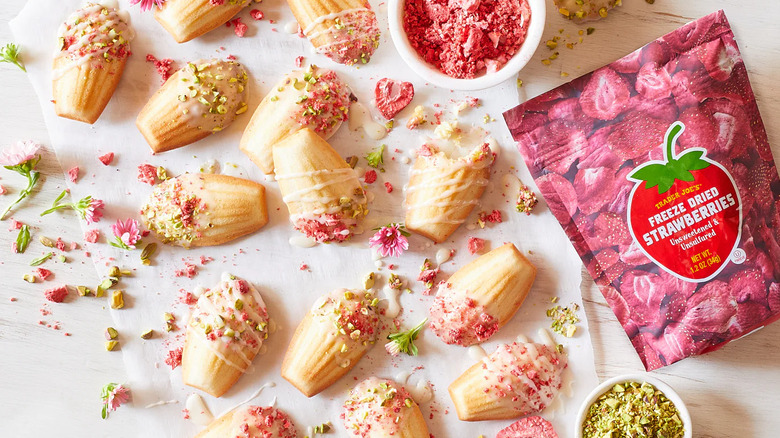 A bag of Trader Joe's Freeze Dried Strawberries on a table with several madeleine cookies scattered on a table top that are coated in crushed freeze dried strawberries and pistachios