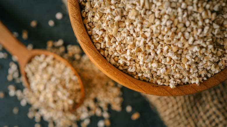 Wooden spoon filled with steel-cut oats, scattered grains on dark table, wooden bowl and burlap background.