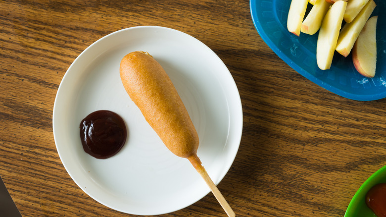 Corn battered hot dog, corn dog, simple kids school lunch beside a bit of ketchup on the plate for dipping