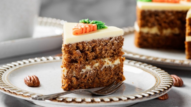a slice of carrot cake with a piped carrot on top. A larger cake with a slice missing sits in the background