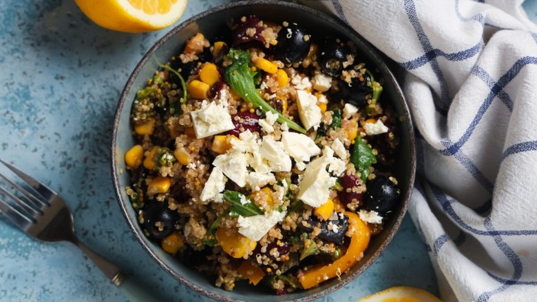a bowl of quinoa with vegetables and feta cheese on a blue counter next to silverware and a cloth