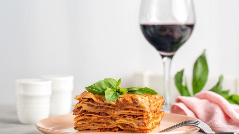 A piece of lasagna with fresh basil on a plate in front of a glass of red wine