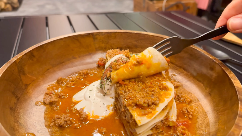 Fork cuts into a piece of taco lasanga in a shallow wooden dish with sour cream and saucy ground beef