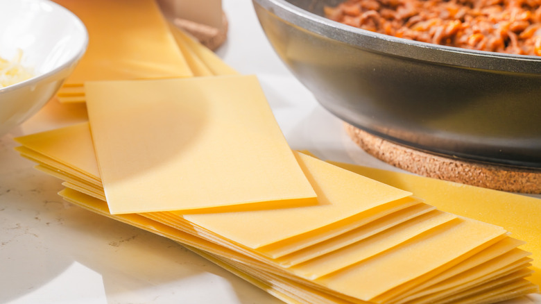 A pile of fresh pasta sheets on white countertop next to bowl of grated cheese and pan with ground meat