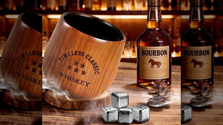 Oak bourbon glass next to whiskey stones, a pinecone, and a bottle of bourbon on a bar counter.