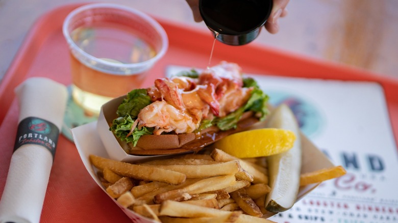 Lobster roll with a side of fries, a pickle slice, and a lemon wedge in a to-go container from Portland Lobster Co.