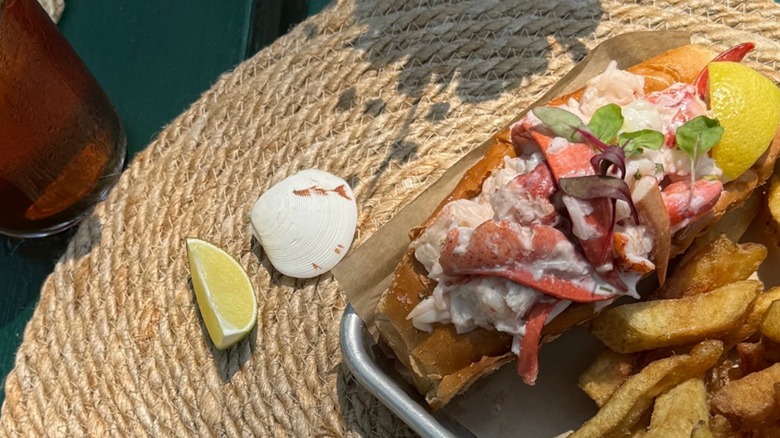 Lobster roll with a side of fries and lemon wedges on a metal tray atop a wicker table Abel's Lobster