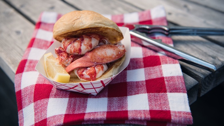 The Clam Shack lobster roll in a to-go container with a lemon wedge on a red plaid napkin on a wooden table