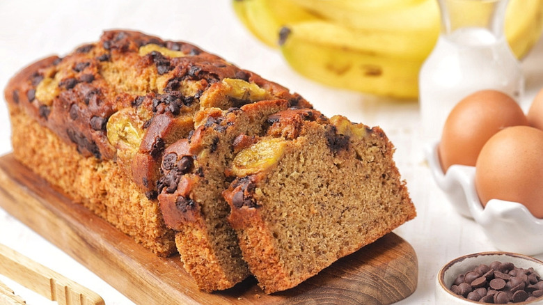 Banana bread sliced on a board surrounded by eggs, chocolate chips, and bananas