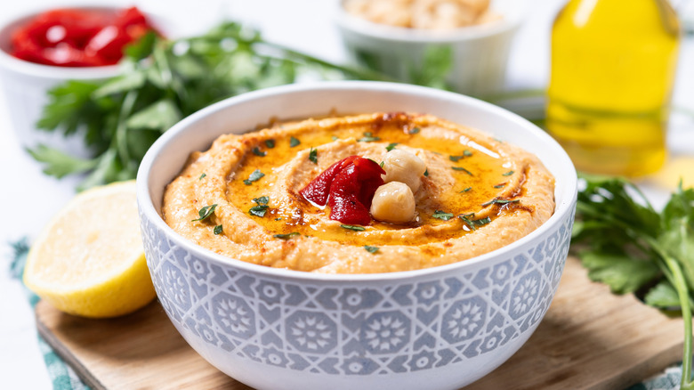 Roasted red pepper hummus in a decorated white bowl in front of herbs and red peppers