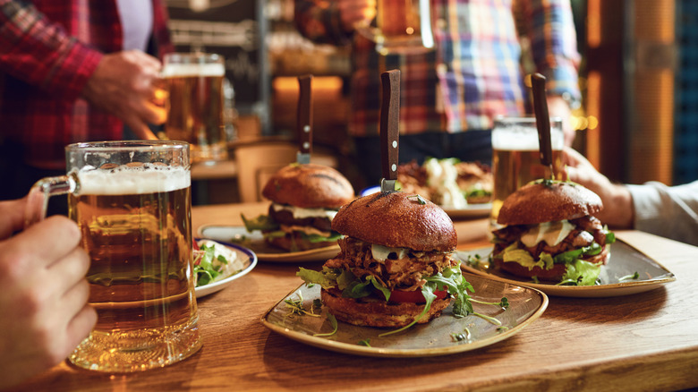 Three burgers on table with men holding beers.
