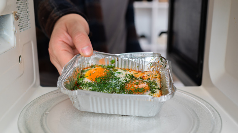 Hands place foil container with unch in microwave to heat.