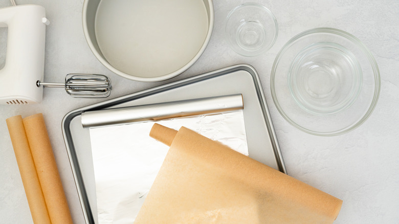 Flat lay composition featuring parchment paper, aluminum foil, a baking pan, mixer, and glass bowls arranged in a close-up view on a light grey background.