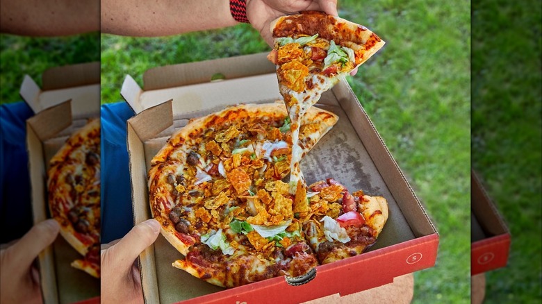 A person holding a box of Casey's taco pizza on their lap and pulling out a slice.