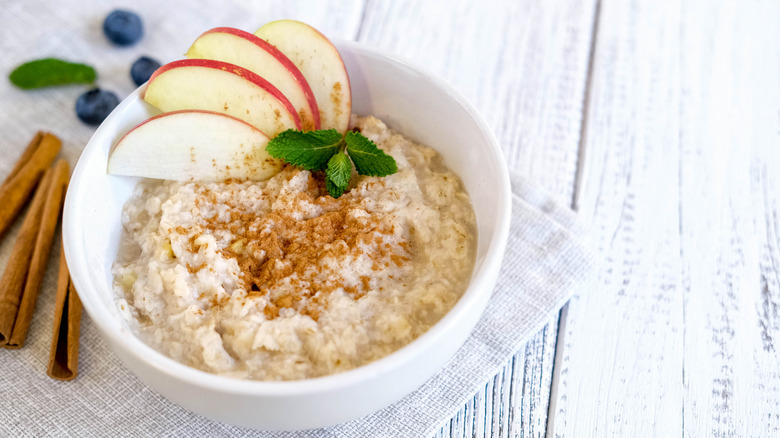 Bowl of oatmeal with apple slices, cinnamon, and mint sprig on white table