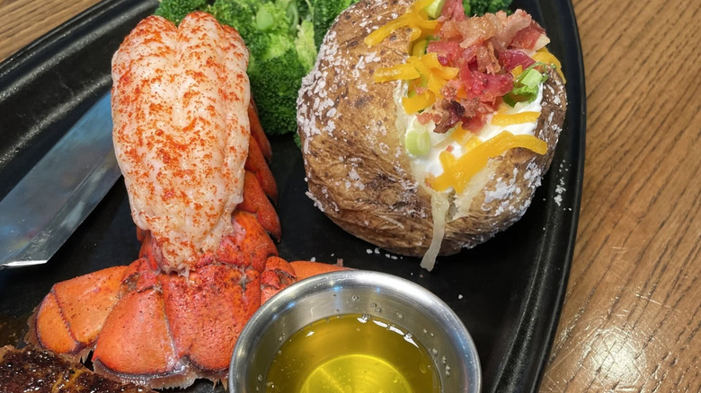 Lobster tail next to a loaded baked potato, butter, and some broccoli and a black plate