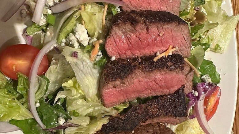 Medium rare steak pieces atop a salad with some cheese, onions, and tomatoes on a white plate