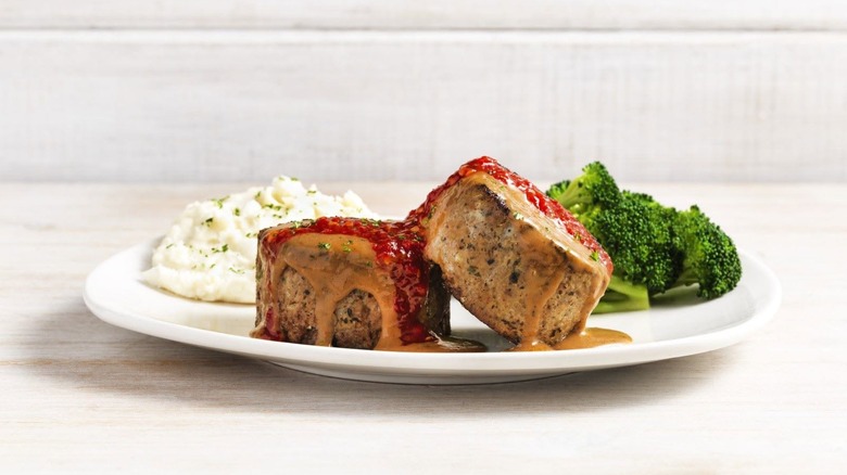 Two pieces of meatloaf covered in sauce on a white plate with broccoli and mashed potatoes