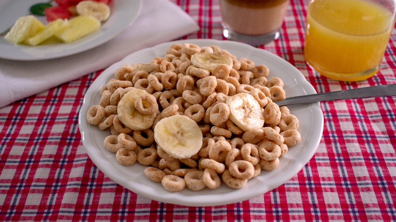 Cheerios topped with banana slices in a white bowl atop a red-checkered tablecloth