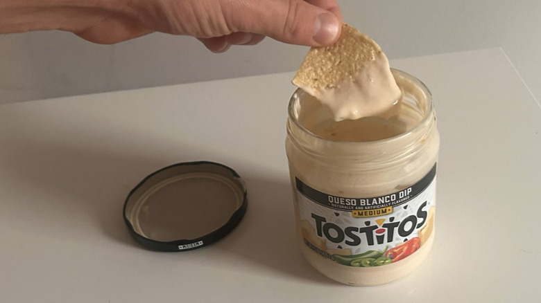 A hand dipping a chip into a jar of tostitos queso blanco