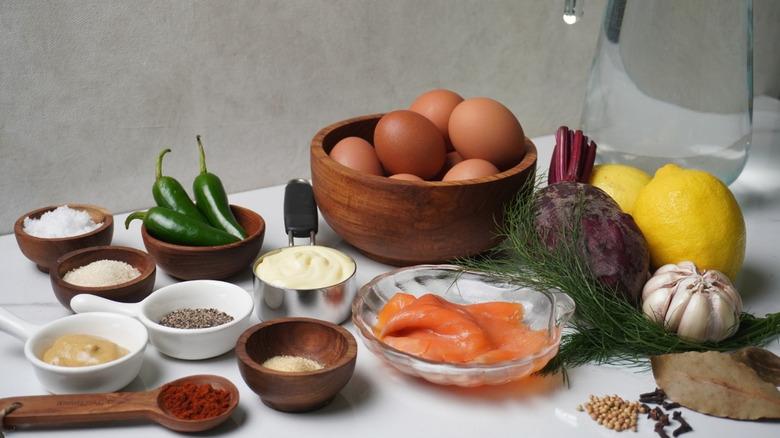 A bowl of eggs on a white countertop with seasonings, mustard, mayo, and fresh ingredients