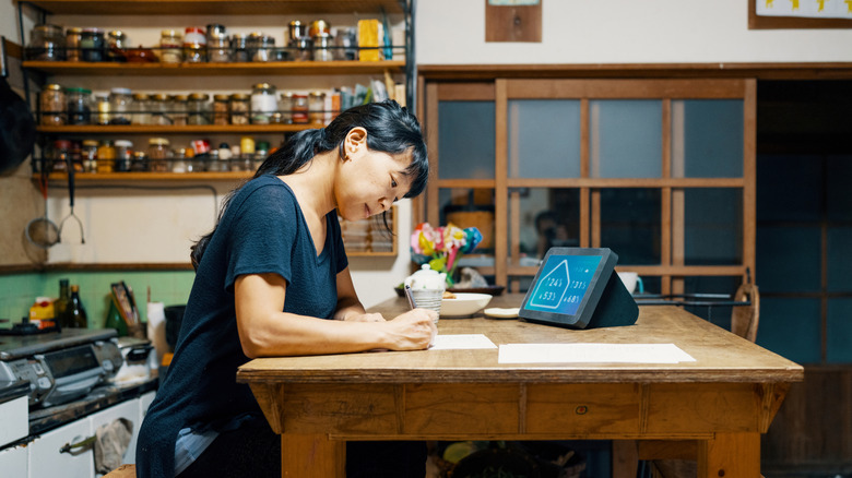 Woman writing at a kitchen table with a wall of spices behind her