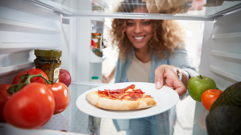 Woman taking a plate of uncovered leftover pizza out of an open fridge