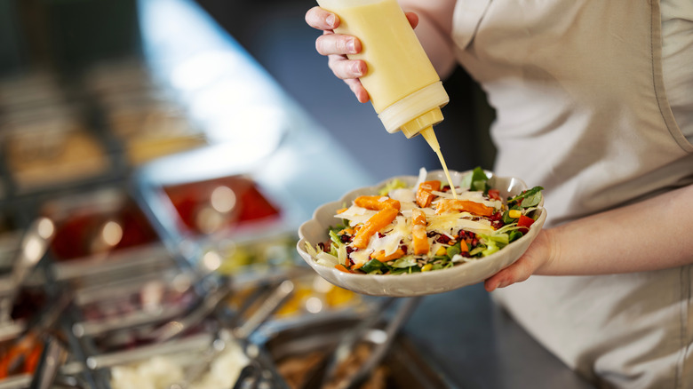 Hand pouring creamy dressing over a bowl of fresh mixed salad with sweet potato and cheese at a buffet