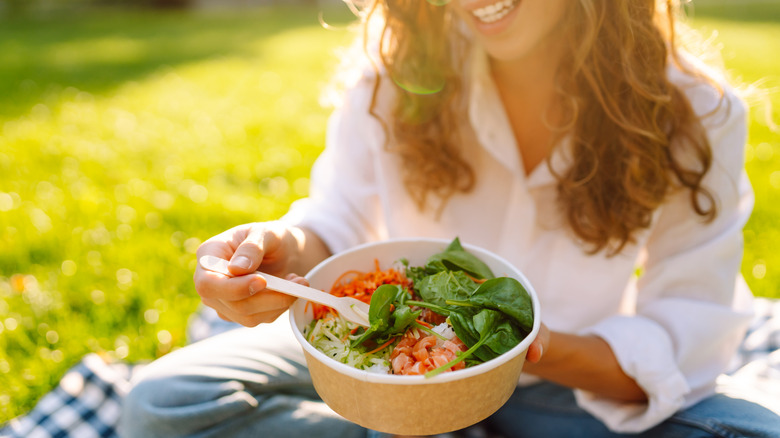 Woman enjoying a salad bowl on a sunny day in a park while sitting on a picnic blanket surrounded by green grass