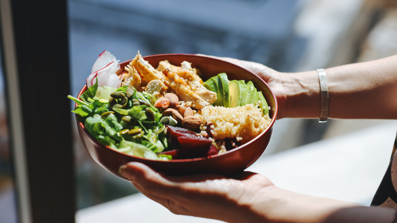 Hands holding a mixed greens salad with beets, avocado, and other vegetables in a bred bowl