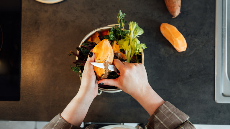 Person peeling a sweet potato over a bowl of vegetables with a paring knife