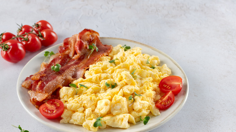 Scrambled eggs on a round, white plate with a golden rim, with bacon and tomatoes, garnished with fresh herbs