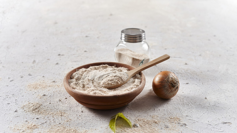 Onion powder in a wood bowl with a wooden spoon, a whole onion on the side, and a shaker of the powder in the background