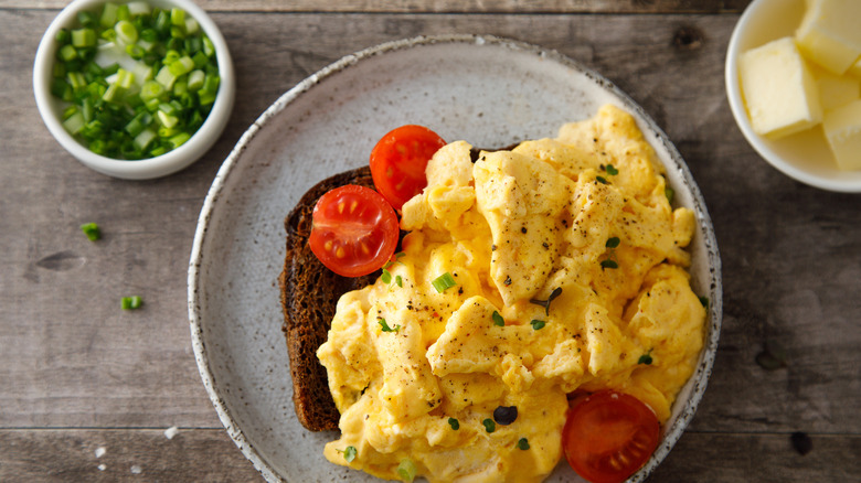 Scrambled eggs and tomatoes on toast sitting on a stoneware plate with ramekins of green onions and butter on the side