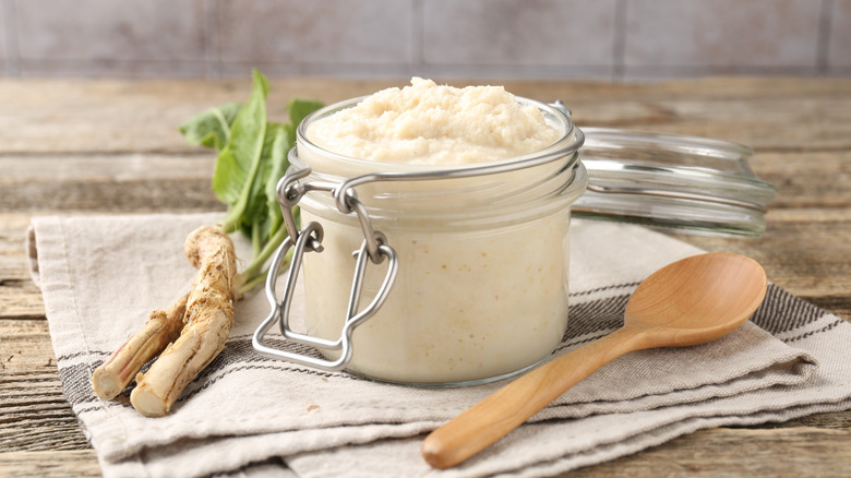 A jar of horseradish sauce, spoon, and roots on a wooden table